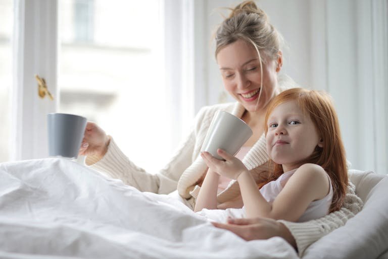 A joyful mother and daughter share a cozy morning with warm drinks in a bright bedroom.