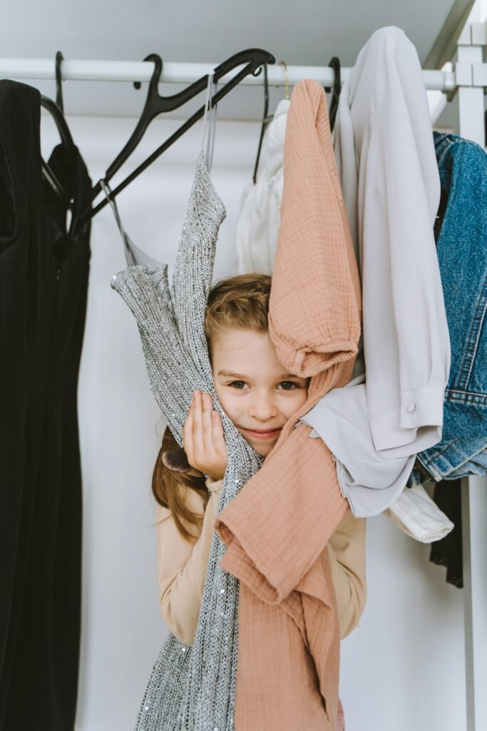 A young girl playfully peeking through hanging clothes in a wardrobe, displaying joy and curiosity.