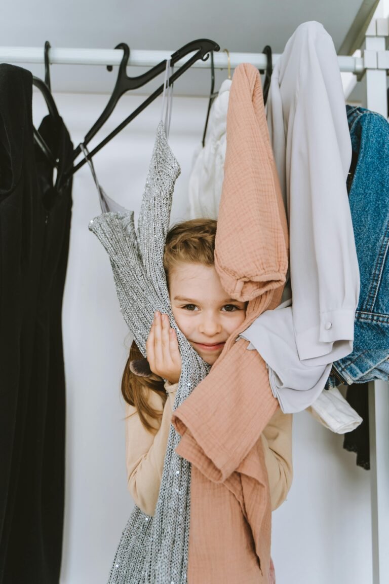 A young girl playfully peeking through hanging clothes in a wardrobe, displaying joy and curiosity.